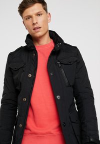 Black padded jacket featuring a high collar, multiple front pockets, and zippered detail. Worn over a coral sweatshirt.