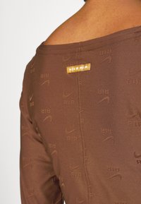 Detailní záběr na hnědé legíny Nike Air s opakovaným vzorem loga a malým žlutým štítkem Nike Air na boku.