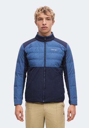 Junger Mann trägt eine blau-navy gestepptes Calvin Klein Jacke mit Frontreißverschluss und beigen Hosen und steht vor einem einfarbigen Hintergrund.
