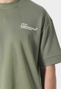 Oliwkowa bawełniana koszulka z krótkim rękawem, z białym nadrukiem tekstu "IRASUTOSTUDIOS" oraz japońskimi znakami na lewej piersi.