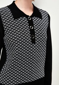 Schwarzes Strickoberteil mit weißem geometrischem Muster, einem Polokragen und drei runden silbernen Knöpfen entlang der Knopfleiste. Glatte Textur.