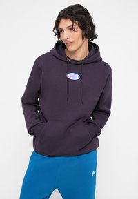Jovem com cabelo encaracolado escuro, vestindo um hoodie Nike roxo escuro e calças de treino Nike azuis brilhantes, com as mãos nos bolsos.