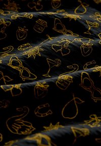 Tissu noir avec broderie métallique dorée présentant des motifs ludiques, notamment des lunettes de soleil, des boissons, des fruits et des motifs de soleil, surface texturée.