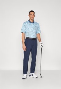 Herre golfantrekk bestående av en lyseblå, kortermet polo med blomstermønster, kombinert med marineblå bukser, et svart belte og hvite golf sko.