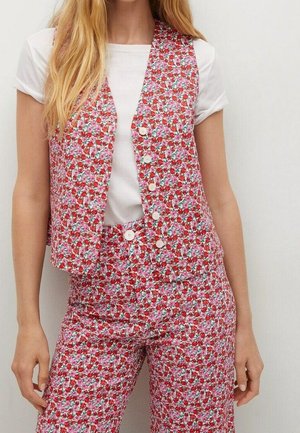 Kobieta ubrana w różowo-czerwony kwiatowy wzorzysty kamizelkę i pasujące spodnie, na którą nałożona jest biała koszulka z krótkim rękawem, stoi na tle jednolitej ściany.