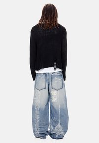 Άτομο με μακριά dreadlocks που φοράει μαύρο πουλόβερ πάνω από άσπρο πουκάμισο και φαρδιά μπλε τζιν με λευκά λουλουδένια και γραφικά σχέδια, γυρισμένο προς τα πίσω.