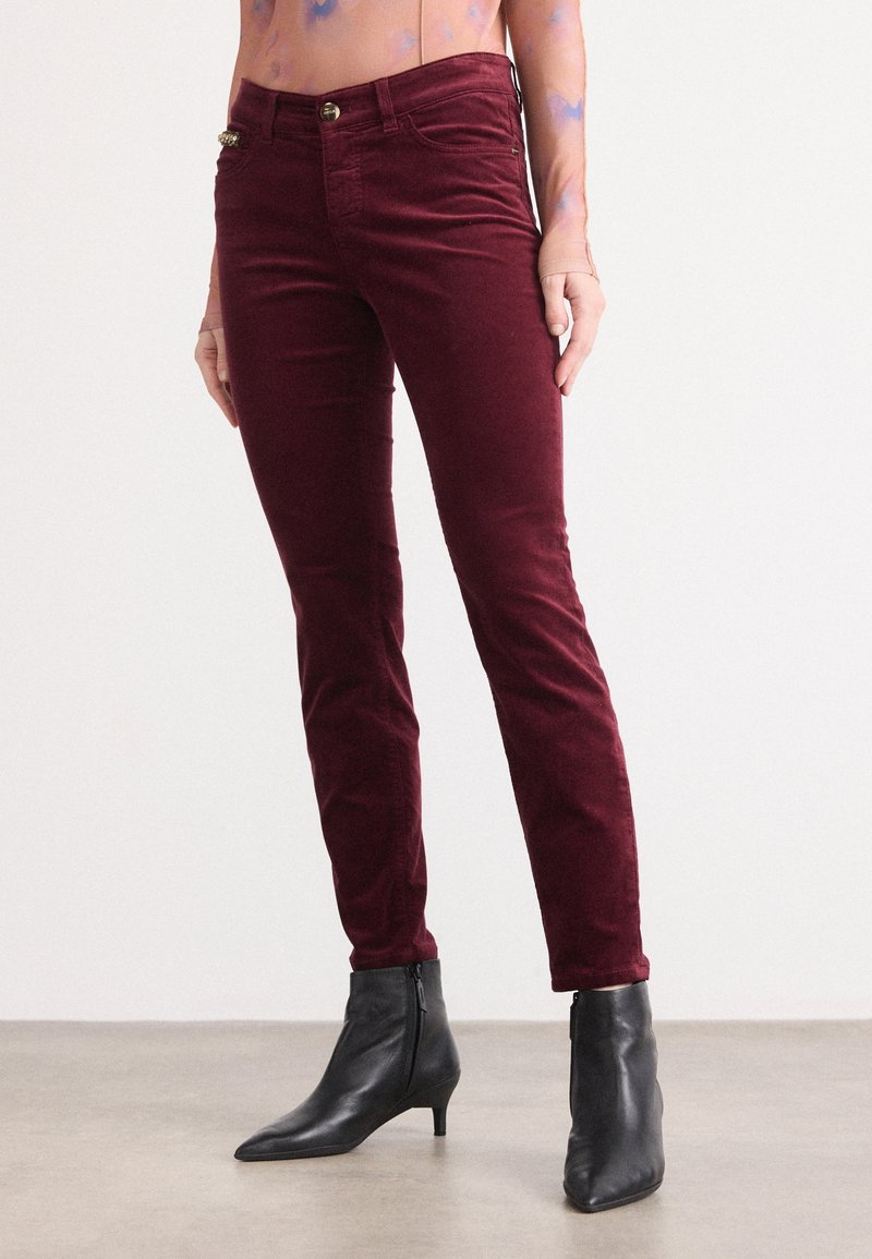Pantalon bordeaux slim-fit porté avec des bottines noires à bout pointu sur une personne debout devant un fond uni.