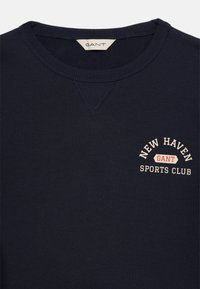 Námořnická mikina s kulatým výstřihem, s logem, které na hrudi zní "New Haven GANT Sports Club" v bílém a červeném textu.