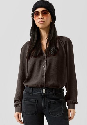 Frau mit langem dunklem Haar, trägt eine schwarze Mütze, orange getönte Sonnenbrille, eine dunkelbraune Bluse und schwarze Jeans mit aufgesetzten Taschen.