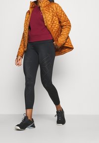 Giacca arancione trapuntata, felpa bordeaux, leggings neri con fantasie a pois e scarpe da ginnastica nere, che mettono in mostra un look sportivo a strati.