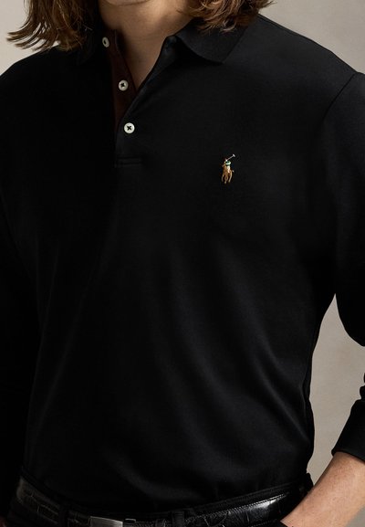Personne portant un polo noir à manches longues avec des boutons blancs et un logo joueur de polo brodé sur la poitrine.