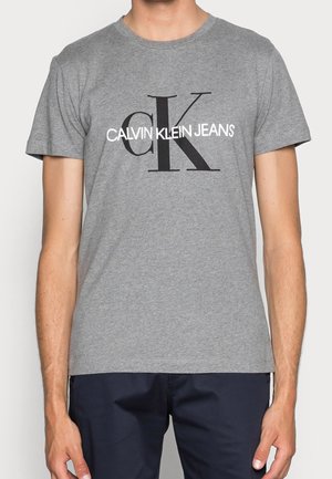 Homme portant un t-shirt gris à manches courtes avec un grand "cK" noir et un texte blanc superposé "CALVIN KLEIN JEANS" sur le devant.