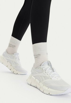 Beine in schwarzen Leggings, weißen Reebok-Socken und weißen Reebok-Sneakers mit dicken, strukturierten Sohlen vor einem einfarbigen Hintergrund.