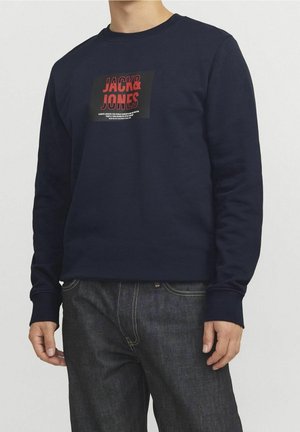 Mężczyzna w granatowej bluzie z czerwono-czarnym logo „JACK & JONES” na piersi oraz ciemnoniebieskich dżinsach na białym tle.