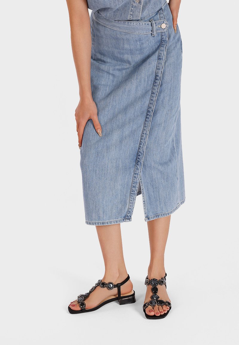 Gonna avvolgente in denim di colore azzurro chiaro, con bottone laterale e spacco. Sandali neri flat con perline decorative e dettaglio della cinghia alla caviglia.