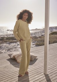 Helles gelbes geripptes Sweatshirt und passende weite Hose, mit entspannter Passform und strukturiertem Oberflächen. Vor einer Küstenlandschaft.