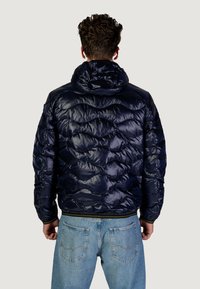 Giacca imbottita blu navy con cappuccio, caratterizzata da un design trapuntato, una texture lucida, polsini elasticizzati e una vita aderente. Abbinata a jeans azzurri chiari.