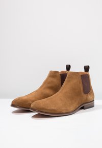 Un paio di stivaletti Chelsea in suede marrone con pannelli laterali elastici scuri e linguette per il tiro, esposti su una superficie bianca.