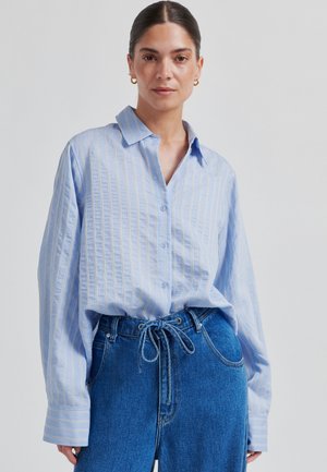 Jasnoniebieska koszula zapinana na guziki, z długimi rękawami i kołnierzykiem. Do tego wysokie, ciemnoniebieskie dżinsy z denimu.