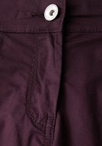 Tessuto di cotone viola scuro con cuciture visibili e chiusura a bottone argentato. Presenta una tasca laterale delineata da cuciture curve.
