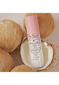 Too Faced Hangover 3-in-1 spray in een roze en witte cilindervormige fles tussen twee natuurlijke kokosnootschalen met een vezelige textuur.