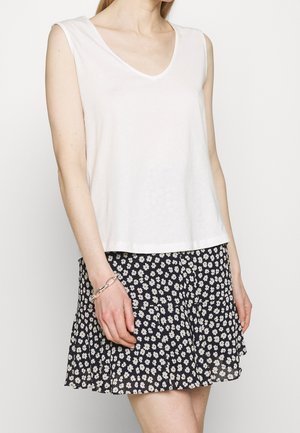 Femme portant un haut blanc sans manches à encolure en V et une jupe noire à motifs floraux blancs, debout avec le bras gauche détendu le long du corps.