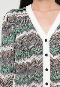 Cardigan țesut în negru, alb și verde cu un model în zigzag. Dispune de un decolteu în V și butoane negre pe partea din față.