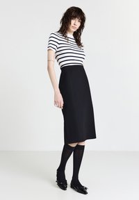 Donna con capelli ricci bagnati, che indossa una gonna nera midi, calze nere fino al ginocchio, scarpe nere e una maglietta bianca a maniche corte con strisce nere.