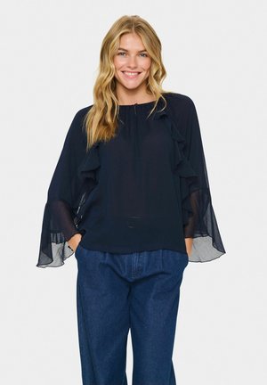 Navy blue blouse made of sheer fabric with ruffled accents along the sleeves, featuring a round neckline and loose fit. Styled with blue trousers.