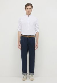 Camicia bianca a maniche lunghe con un design senza colletto, abbinata a pantaloni blu scuro e una cintura marrone. Scarpe beige casual completano l'outfit.