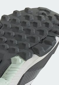 Semelle de chaussure en caoutchouc noir avec des crampons hexagonaux et des motifs texturés, tige en mesh gris et blanc et accents de coussinets visibles.