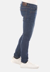 Niebieskie dżinsy w slim fit, z kontrastującymi pomarańczowymi przeszyciami i skórzaną łatą na pasie. Widok z boku ukazuje szczegóły kieszeni.