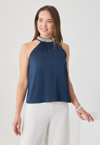 Sötétkék nyakpántos felső sima anyagból, redőzött nyakkivágással és gyöngyös gallérral. Fehér, bőszárú nadrággal párosítva.