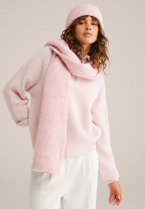 Maglione rosa chiaro lavorato a maglia in materiale peloso, abbinato a un berretto e una sciarpa a costine coordinati. I pantaloni bianchi a costine completano il look.