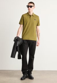 Camisa polo de color verde oliva con diseño de cuello, detalle de etiqueta negra, combinada con pantalones negros y sosteniendo una chaqueta de cuero negra. Botas negras.