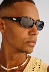 Κοντινό πλάνο ενός άνδρα που φοράει μαύρα ορθογώνια γυαλιά ηλίου, κρεμ πλεκτό μπλουζάκι και περιδέραιο με χάντρες, στρέφοντας το πρόσωπό του προς τα δεξιά σε απλό φόντο.