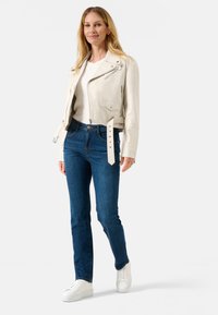 Weiße Lederbikerjacke mit silbernen Akzenten, Frontreißverschluss und seitlichen Taschen, kombiniert mit blauen Jeans und weißen Sneakern.