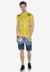 Gelbes Kurzarm-T-Shirt mit bedrucktem Text und Mustern; kombiniert mit blauen Jeansshorts mit Taschen und einem Farbverlauf-Design.