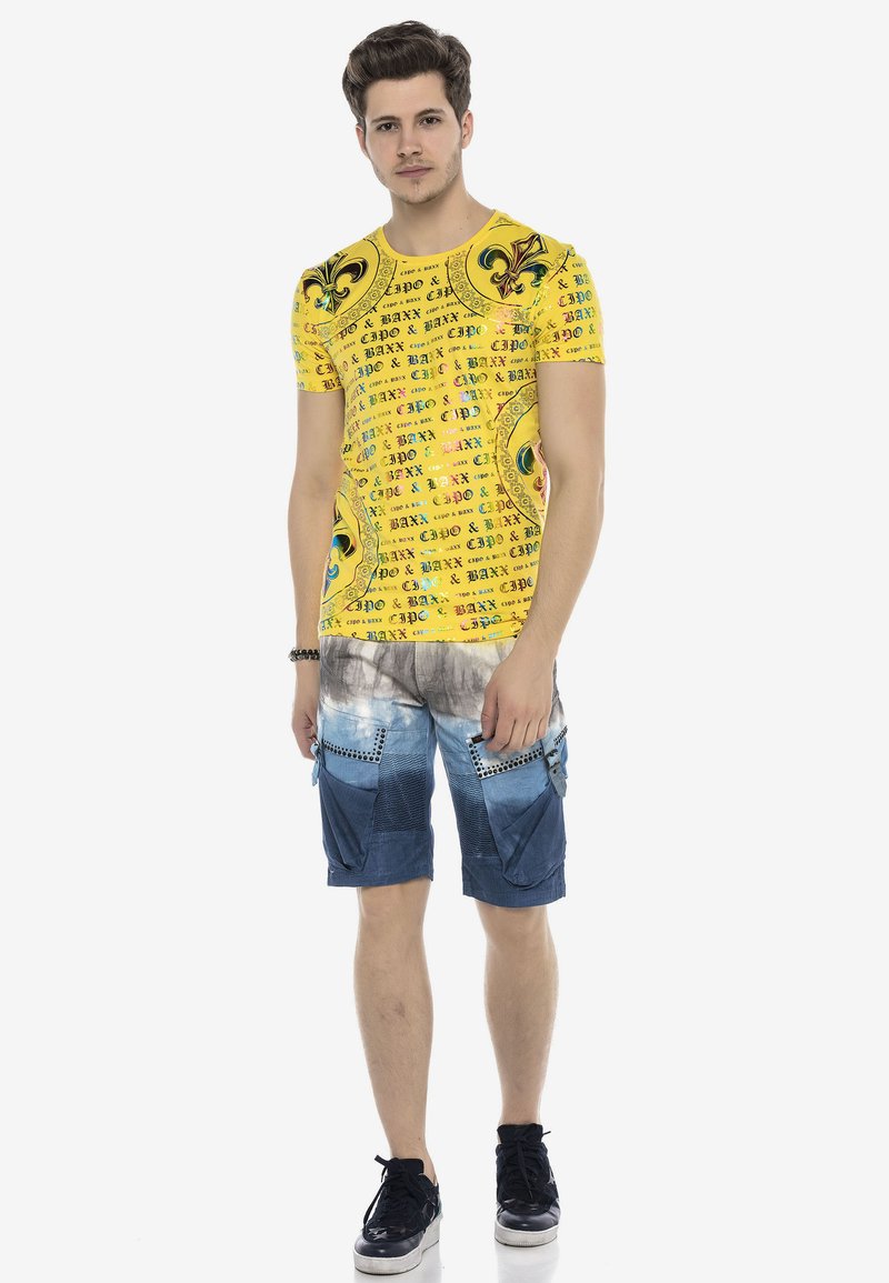 Gelbes Kurzarm-T-Shirt mit bedrucktem Text und Mustern; kombiniert mit blauen Jeansshorts mit Taschen und einem Farbverlauf-Design.