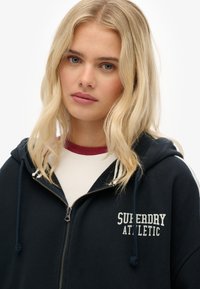 Sudadera con cremallera de color navy con un logotipo blanco de "SUPERDRY ATHLETIC". Tiene capucha, cordones y una textura suave. Llevada sobre una camisa crema con acentos en color burdeos.
