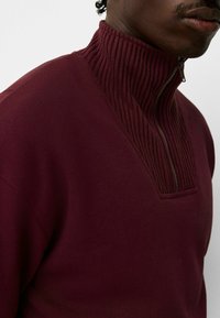 Jersey burdeos con cuello alto de canalé y media cremallera. Tejido suave con un diseño ajustado y detalles texturizados en el escote.