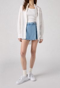 Chemise blanche à manches longues sur un débardeur blanc ajusté, assortie d'une jupe en jean bleu clair et de baskets blanches avec des chaussettes couleur crème.