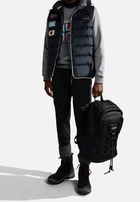 Chaleco de plumas con banderas, sudadera gris, pantalones oscuros, botas negras y una mochila negra con correas. Las características incluyen un diseño acolchado y bolsillos laterales.