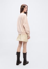 Γυναίκα με μακριά καστανά μαλλιά που φοράει ένα φαρδύ ανοιχτό ροζ φούτερ, μπεζ μίνι φούστα και μαύρα μπότες μέχρι το γόνατο με αγκράφες, στέκεται μπροστά σε μια απλή φόντο.