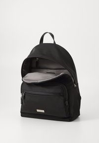 Mochila de nylon negra con parte superior redondeada, interior gris, bolsillo delantero con cremallera y asa superior para llevar. Incluye correas ajustables.