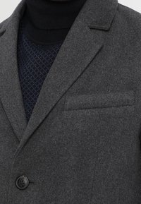 Blazer di lana grigia con rever a incavo, chiusura con un solo bottone e taschino sul petto, indossato sopra un dolcevita blu navy con motivo a fantasia.