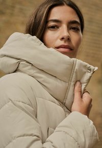 Veste doudoune beige avec un col haut, texture matelassée et détails de boutons. Vue rapprochée se concentrant sur le col et la zone des épaules.