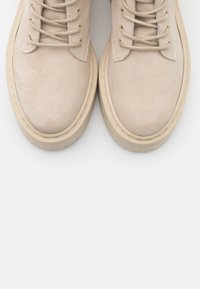 Bottines beiges claires en daim avec un design à lacets et une semelle en caoutchouc épaisse. Elles présentent une texture lisse et des détails de couture contrastants.