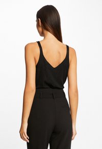 Haut noir sans dos avec des bretelles fines, présentant un décolleté en V et une texture légèrement scintillante, associé à un pantalon noir taille haute.