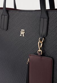 Czarna torba na ramię z wzorem, o teksturowanej powierzchni, z złotym akcentem logo oraz burgundową metką. Posiada podwójne uchwyty i metalowe zapięcia.